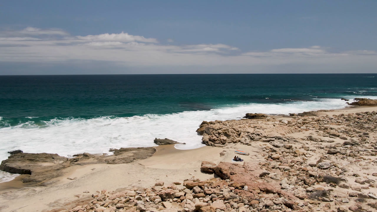 Laguna Beach rocky coast seen from above, with foam spreading along flat rock ledges, a small sandy beach below, open ocean beyond, and small distant people grouped near a bright object on the sand.