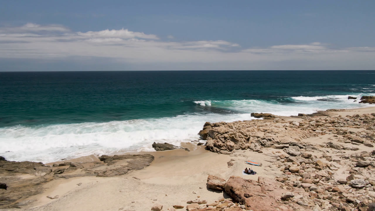 Laguna Beach coastline with a broad band of whitewater crossing the midground, tan rocks and sand below, and small people still visible near the surfboard on the lower beach.