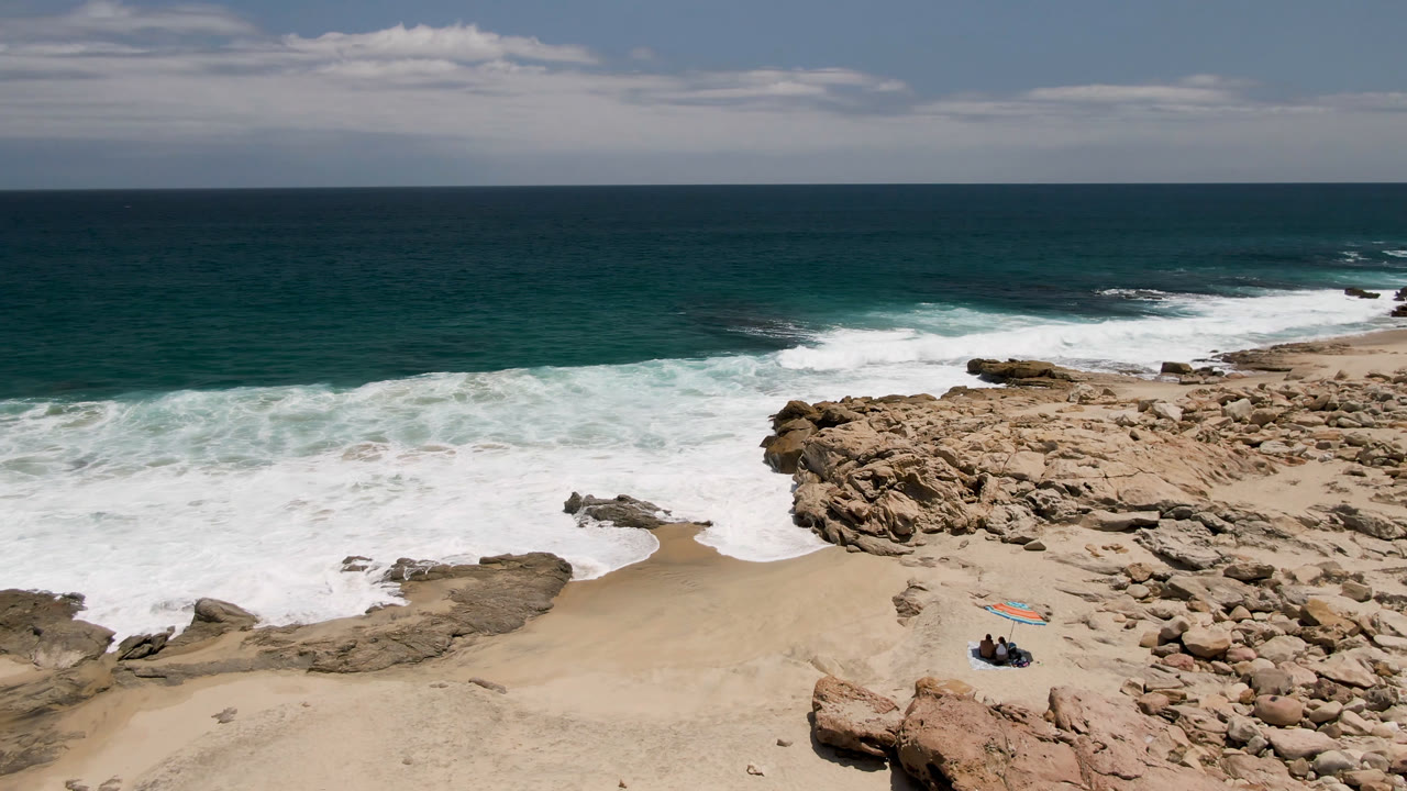 At 0:01, Laguna Beach surf spreads farther across the sand while the distant people and bright beach gear remain near the lower beach below the rock point.
