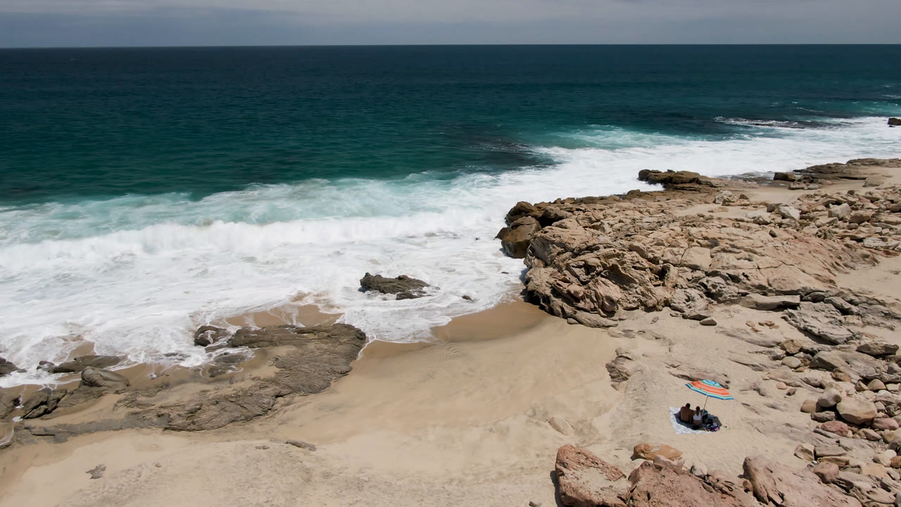 Aerial view of rocky beach, surf, and surfers with boards on sand.