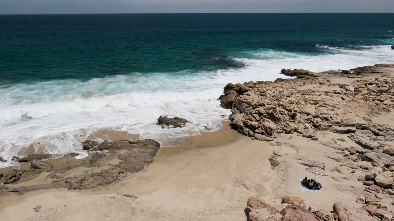 Laguna Beach shoreline seen from above as a foamy wave spreads across the sand, with surfers and colorful boards still visible near the lower right foreground.
