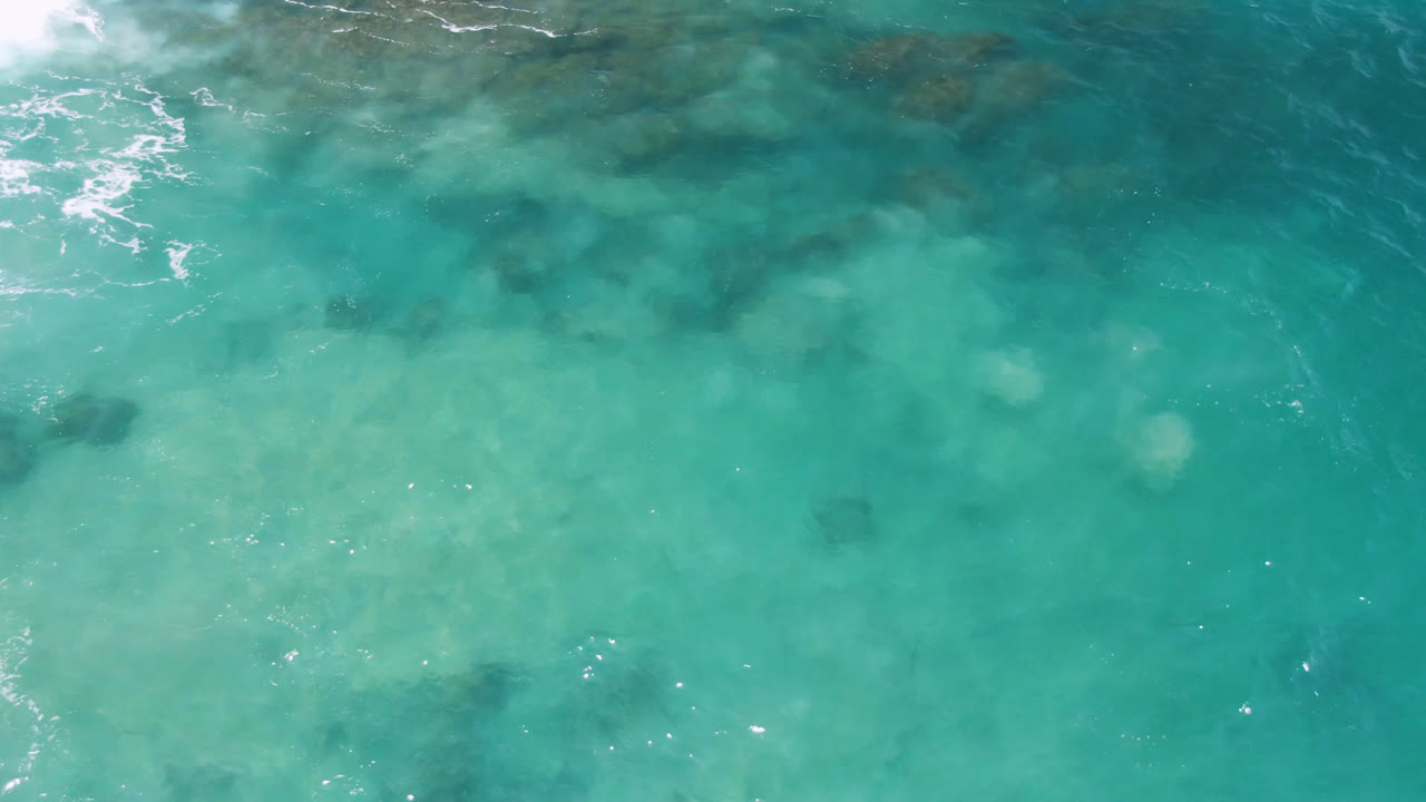 Overhead view of turquoise water at Laguna Beach, Baja California Sur, with pale foam along the upper left edge, darker underwater patches, and no visible people.
