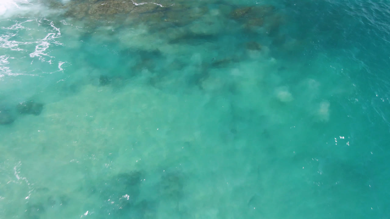 Aerial frame over Laguna Beach, Baja California Sur, showing milky turquoise shallows, soft wave foam near the top edge, darker underwater forms, and no visible people.