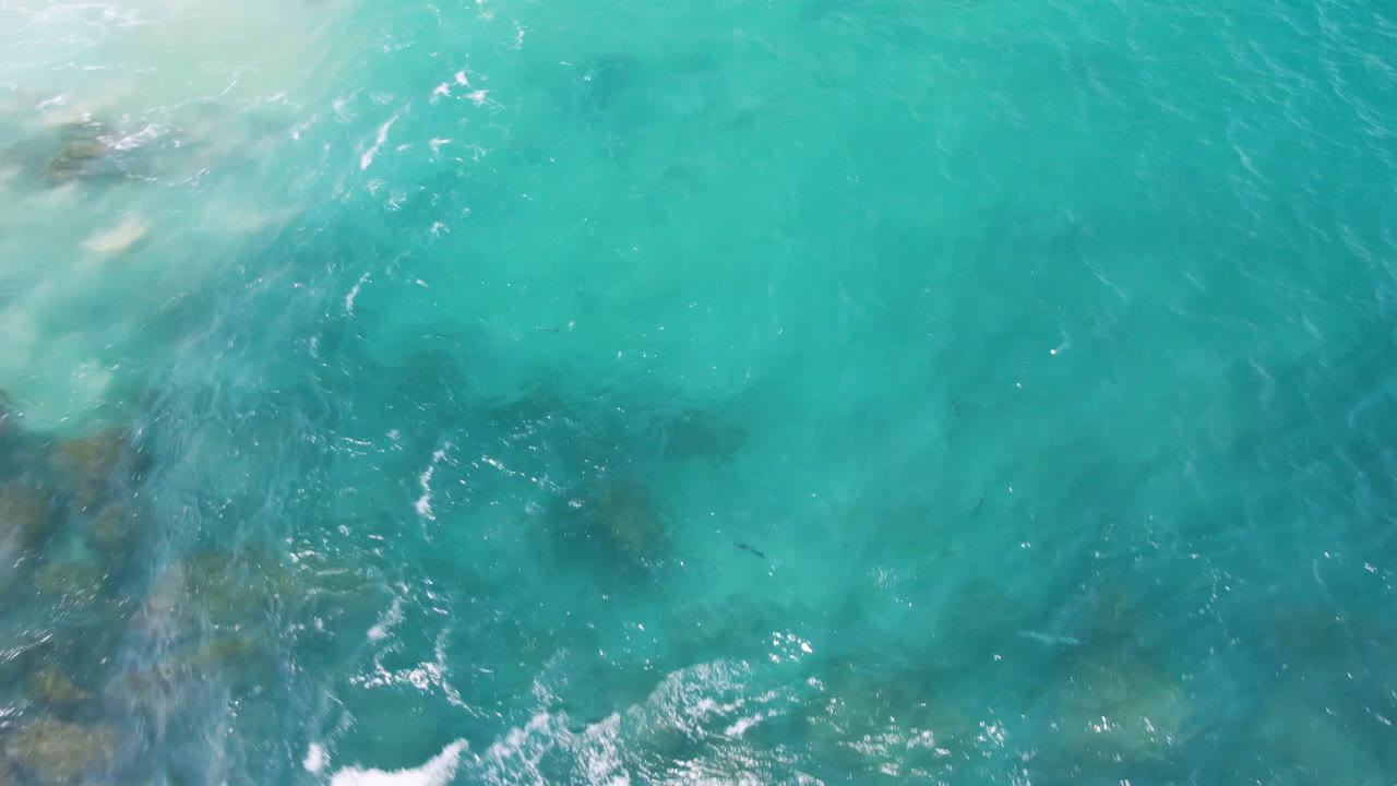 Overhead view of turquoise water at Laguna Beach, Baja California Sur, with faint dark submerged rocks near the left side and no visible people.