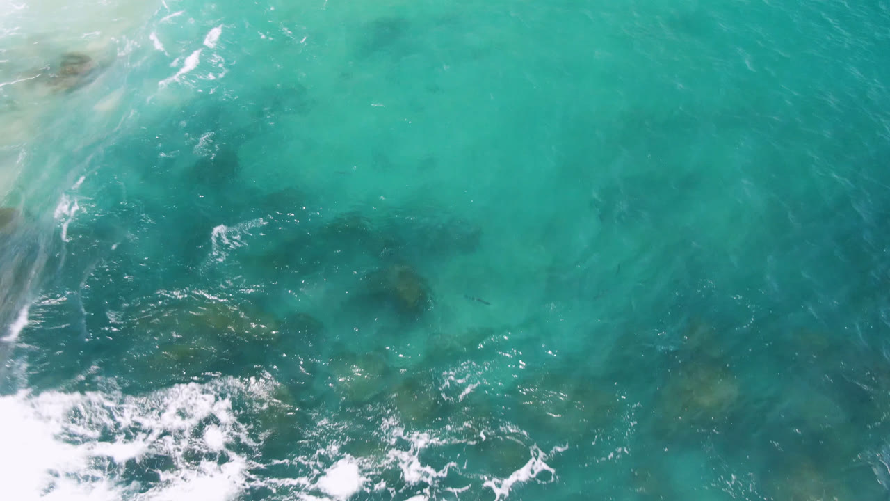 Overhead Laguna Beach water frame with white foam entering from the lower left, darker rock patches below the surface, and no visible people.