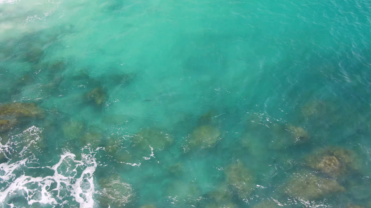 Overhead view of turquoise ocean at Laguna Beach, Baja California Sur, with submerged rock patches, white foam entering from the lower left, and no visible people.