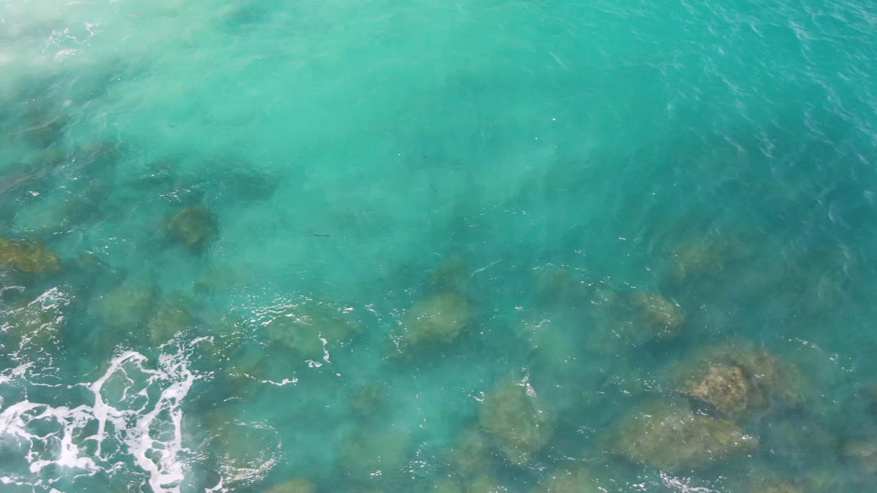 Aerial frame over Laguna Beach turquoise water with darker underwater rock forms and a curling white foam patch near the lower center, with no visible people.