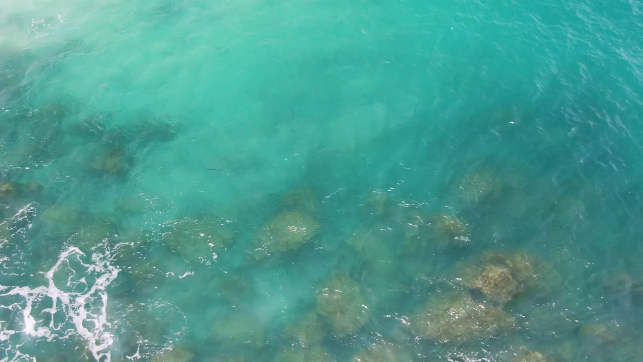 Turquoise ocean surface with scattered submerged rocks and foam near the lower left.