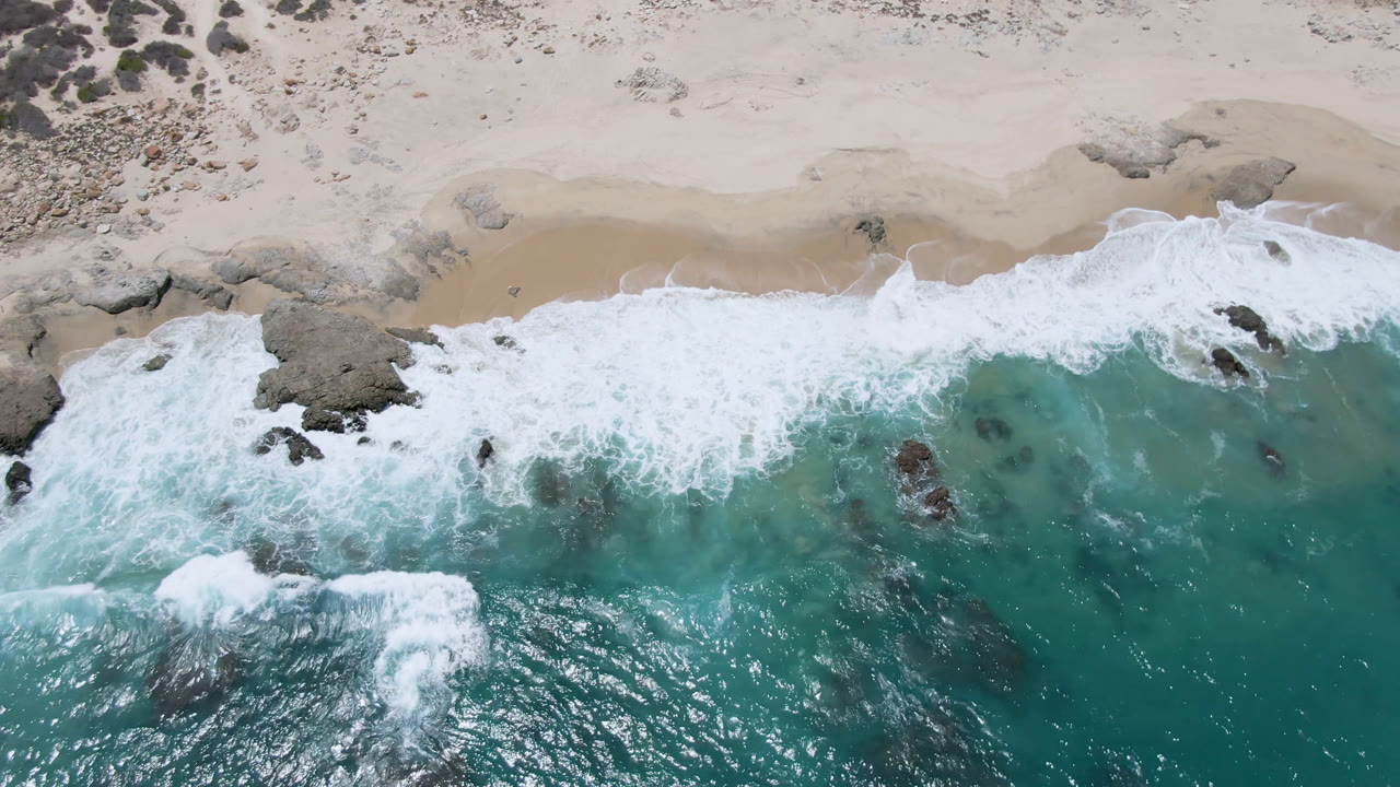 Overhead view of Laguna Beach, Baja California Sur, Mexico, with turquoise water, white surf, pale sand, rock outcrops, and no visible people at the start of the clip.