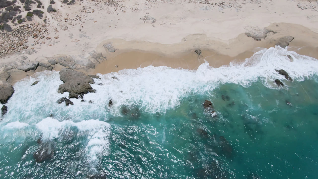 Aerial frame over Laguna Beach, Baja California Sur, Mexico, where a wide band of white foam pushes across turquoise water toward pale sand, with no visible people.