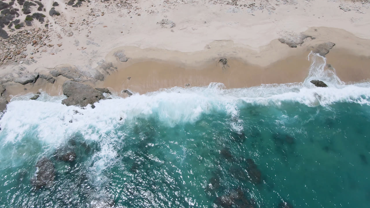 Overhead view of Laguna Beach, Baja California Sur, with turquoise water breaking onto pale sand, dark rocks in the surf, scrub above the beach, and no visible people.