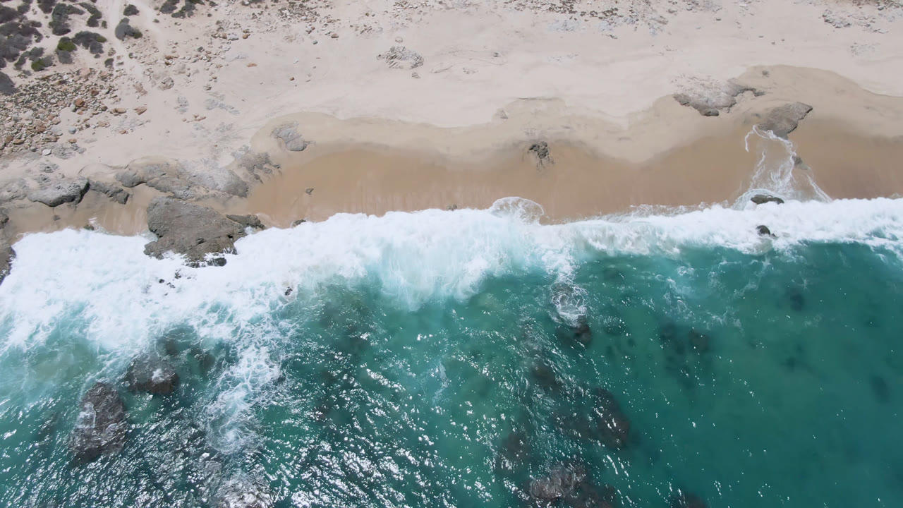 A white wave line spreads across the Laguna Beach, Baja California Sur shoreline, with teal water below, pale sand above, scattered rocks, and no visible people.
