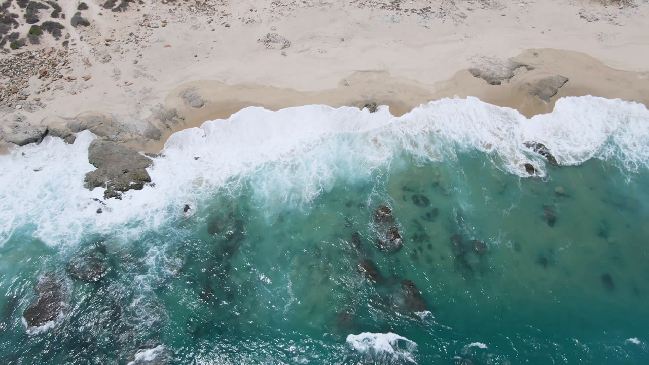 Top-down aerial frame of Laguna Beach, Baja California Sur, showing white surf crossing turquoise water toward pale sand, dark rocks below the surface, scrub along the beach edge, and no visible people.