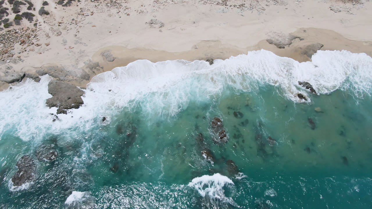 Overhead view of the Laguna Beach shoreline in Baja California Sur, with a broad white wave line spreading over green-blue water, submerged rocks in the lower half, dry sand above, and no visible people.