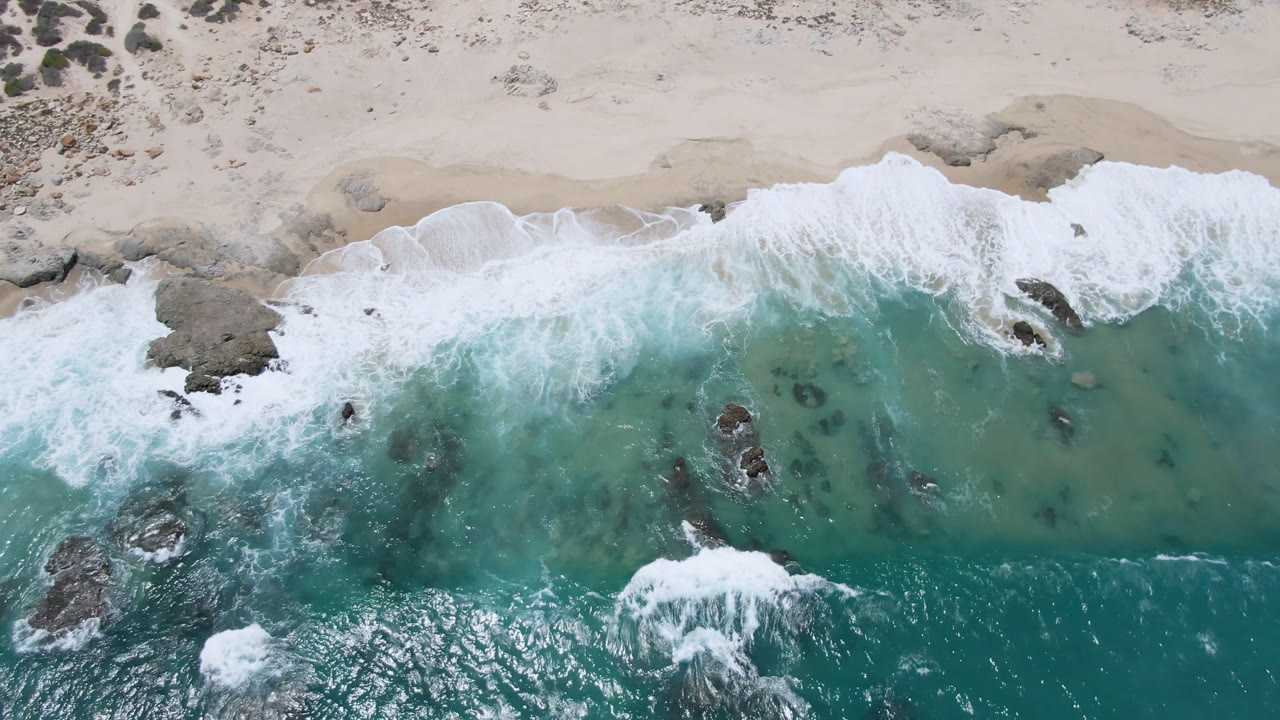 Top-down beach view with teal water, foam, sand, and rocks.