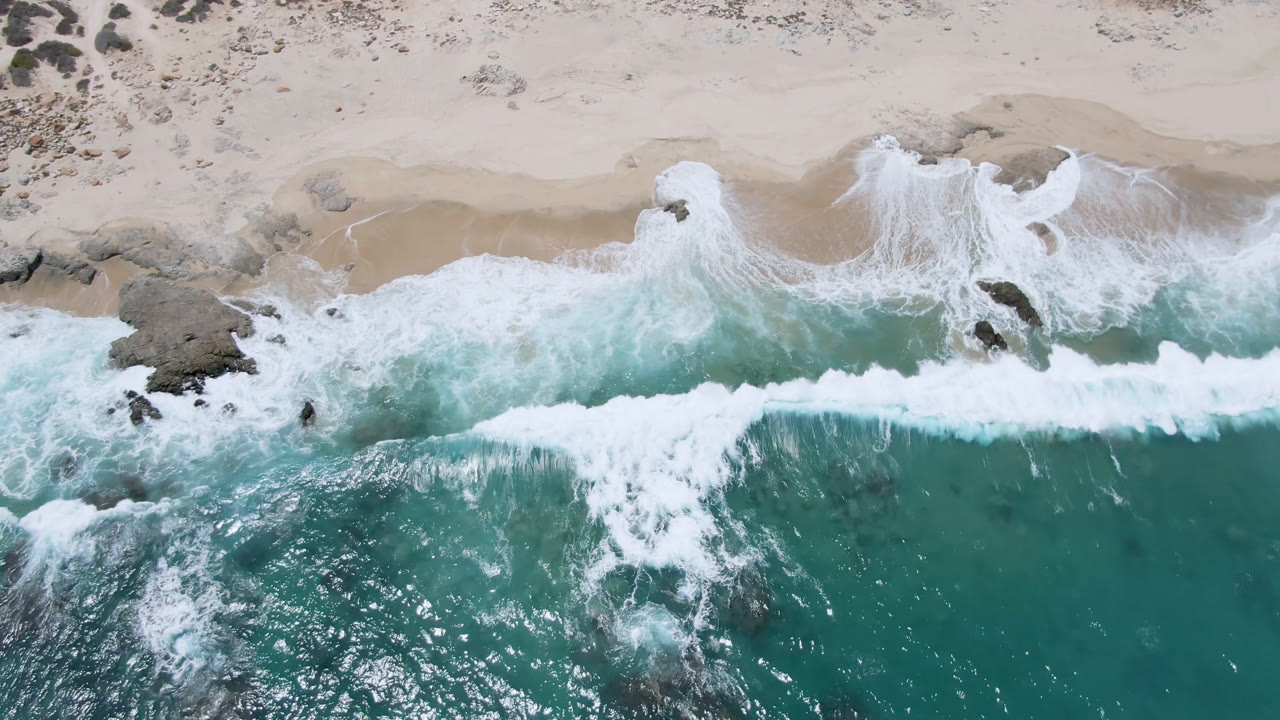 Straight-down aerial view of Laguna Beach in Baja California Sur, with turquoise water, white surf, pale sand, scattered rocks, and no visible people at the start of the clip.