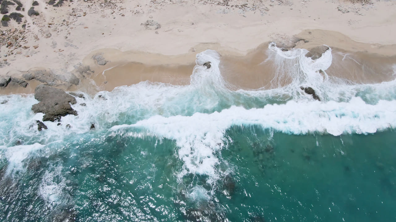 Aerial frame over Laguna Beach in Baja California Sur showing a fresh white wave spread across the shoreline, rocks near the sand, and no visible people.