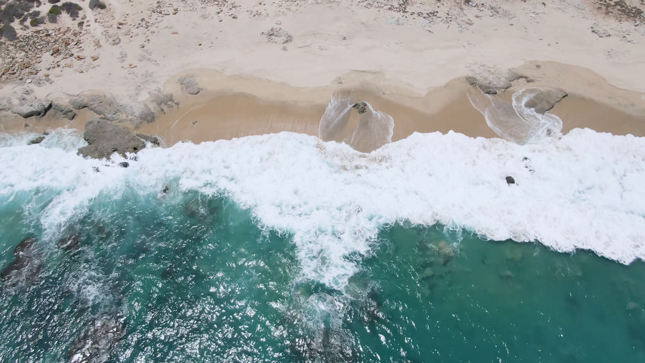 Overhead view of Laguna Beach in Baja California Sur with teal ocean, white surf, pale sand, scattered rocks, and no visible people at the opening moment.