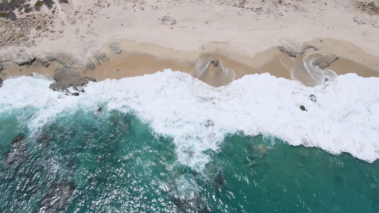 At 0:01, Laguna Beach surf pushes wider across the sand, with teal water below, dark rocks near the break, pale beach above, and no visible people.