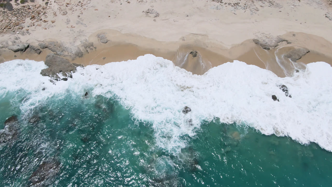 Overhead beach view with a broad band of white surf and exposed dark rocks.