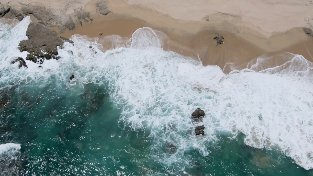 Overhead surf washing onto sandy beach with dark rocks