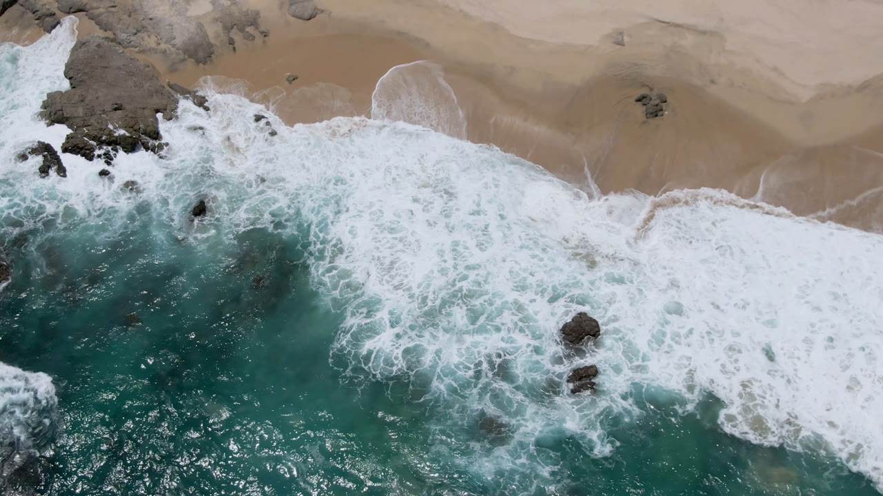 Aerial frame over Laguna Beach, Baja California Sur, with foamy water curling around several dark rocks below a smooth strip of sand and no visible people.