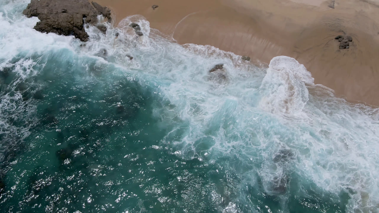 Aerial frame over Laguna Beach, Baja California Sur, where a thicker band of white foam pushes onto the sandy beach beside dark rocks, no visible people.