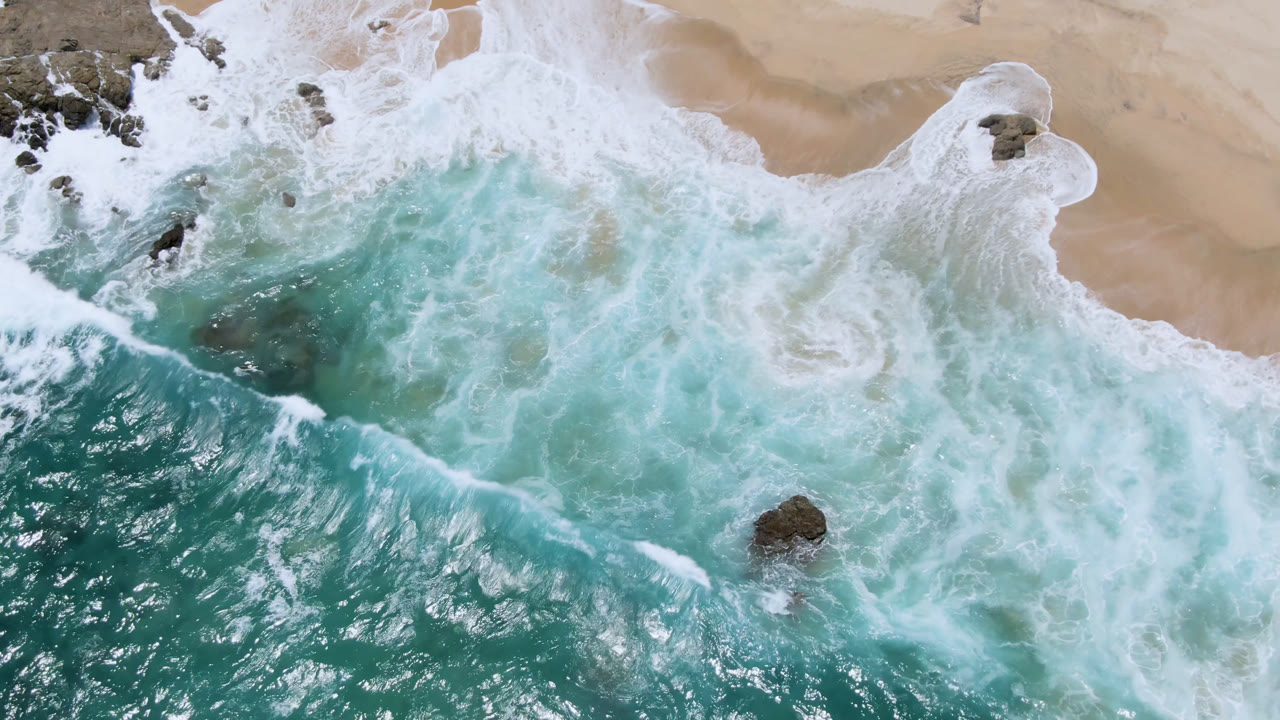 Overhead view at Laguna Beach, Baja California Sur, with turquoise water, white foam, tan sand, scattered rocks, and no visible people at the start of the clip.