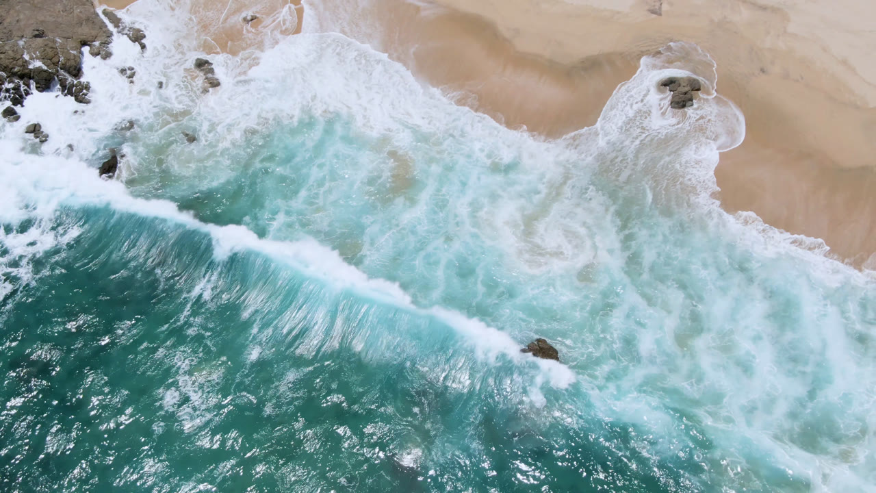 Aerial frame from Laguna Beach, Baja California Sur, showing a bright white wave crest moving across turquoise water toward tan sand, with no visible people.