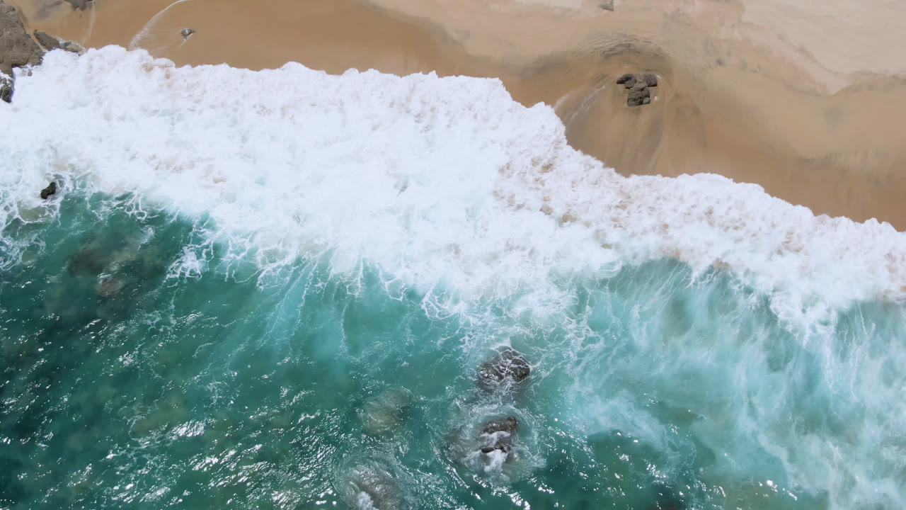 Aerial frame over Laguna Beach, Baja California Sur, with a wide white band of breaking foam, teal water below, sand above, and no visible people.