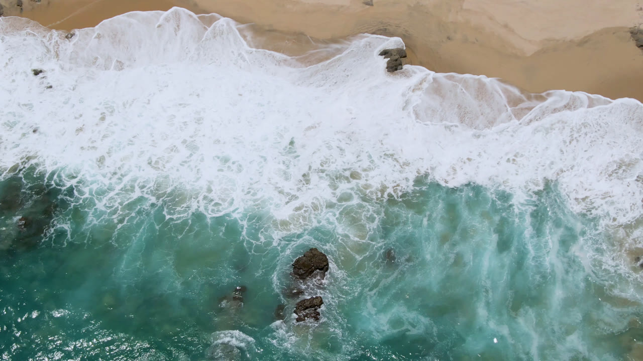 Overhead view of Laguna Beach in Baja California Sur with turquoise water, white seafoam, tan sand, dark rocks, and no visible people at the start of the clip.