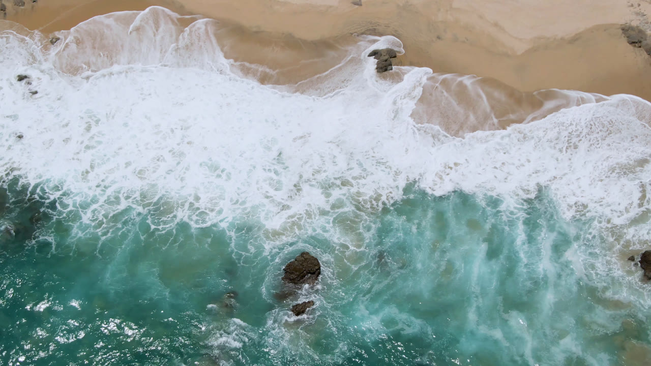 Aerial frame over Laguna Beach in Baja California Sur showing a broad band of white foam washing over tan sand with turquoise water below and no visible people.