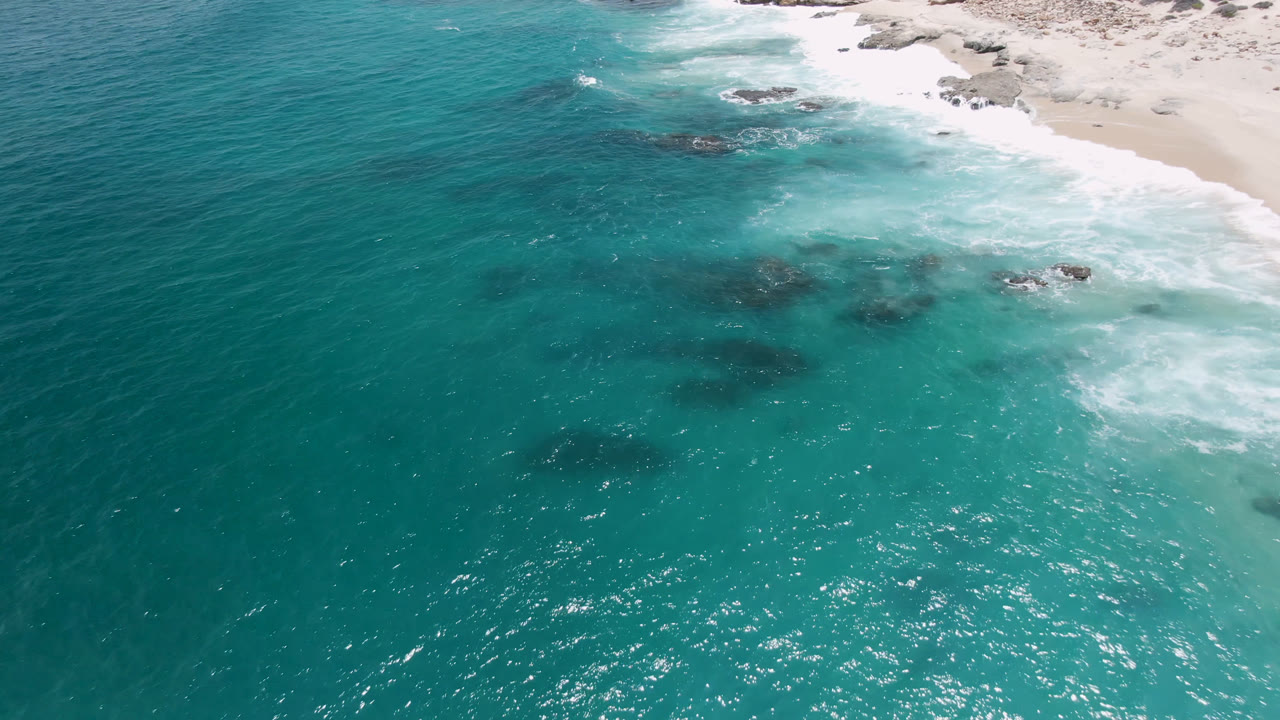 Aerial frame from Laguna Beach, Baja California Sur, showing turquoise ocean, pale sand, white surf, scattered rocks, and no visible people at the start of the clip.