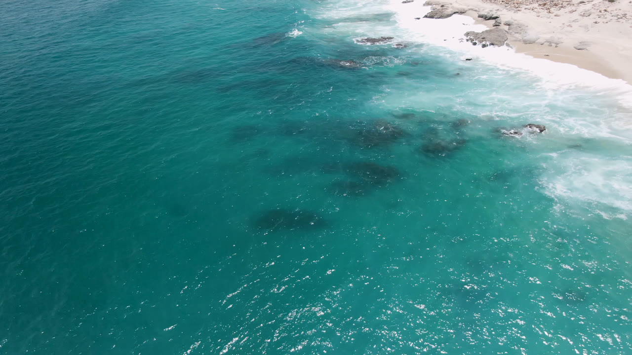 Aerial view over Laguna Beach, Baja California Sur, with rippled turquoise water in the foreground, surf washing the beach near the top, rocks offshore, and no visible people.