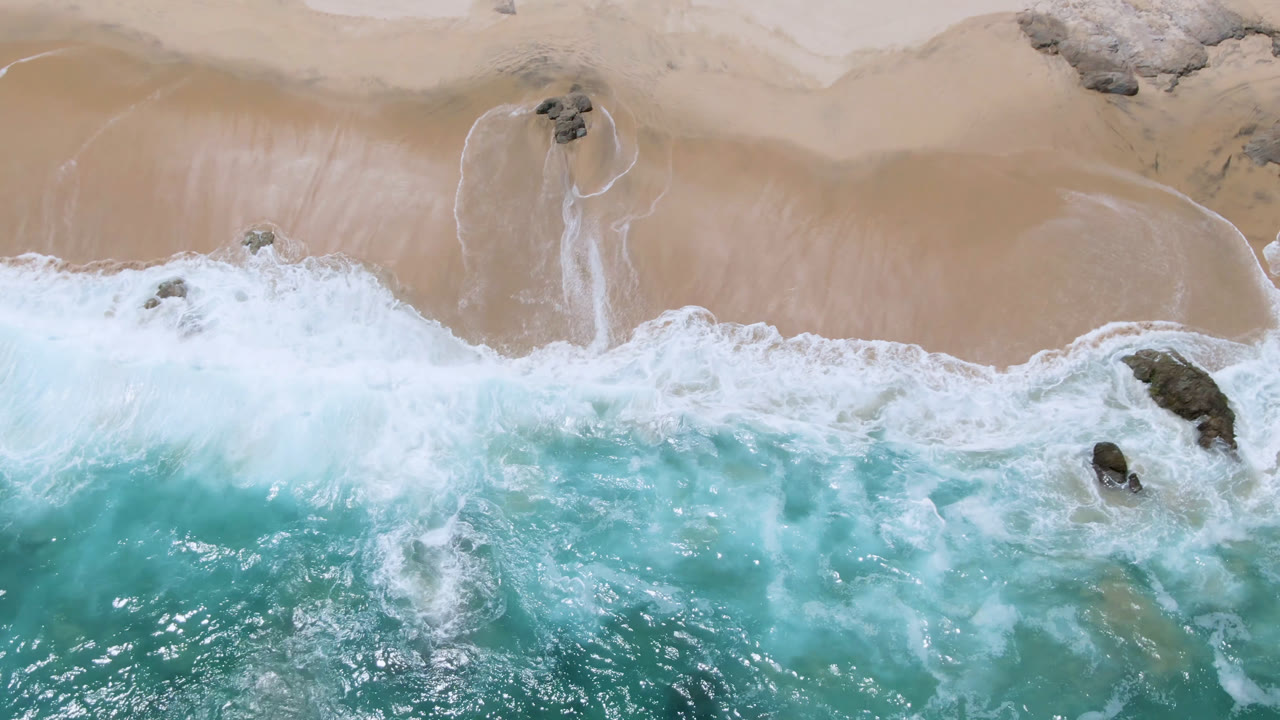 Overhead view of Laguna Beach in Baja California Sur with turquoise water, white foam, tan sand, scattered rocks, and no visible people at the start of the clip.