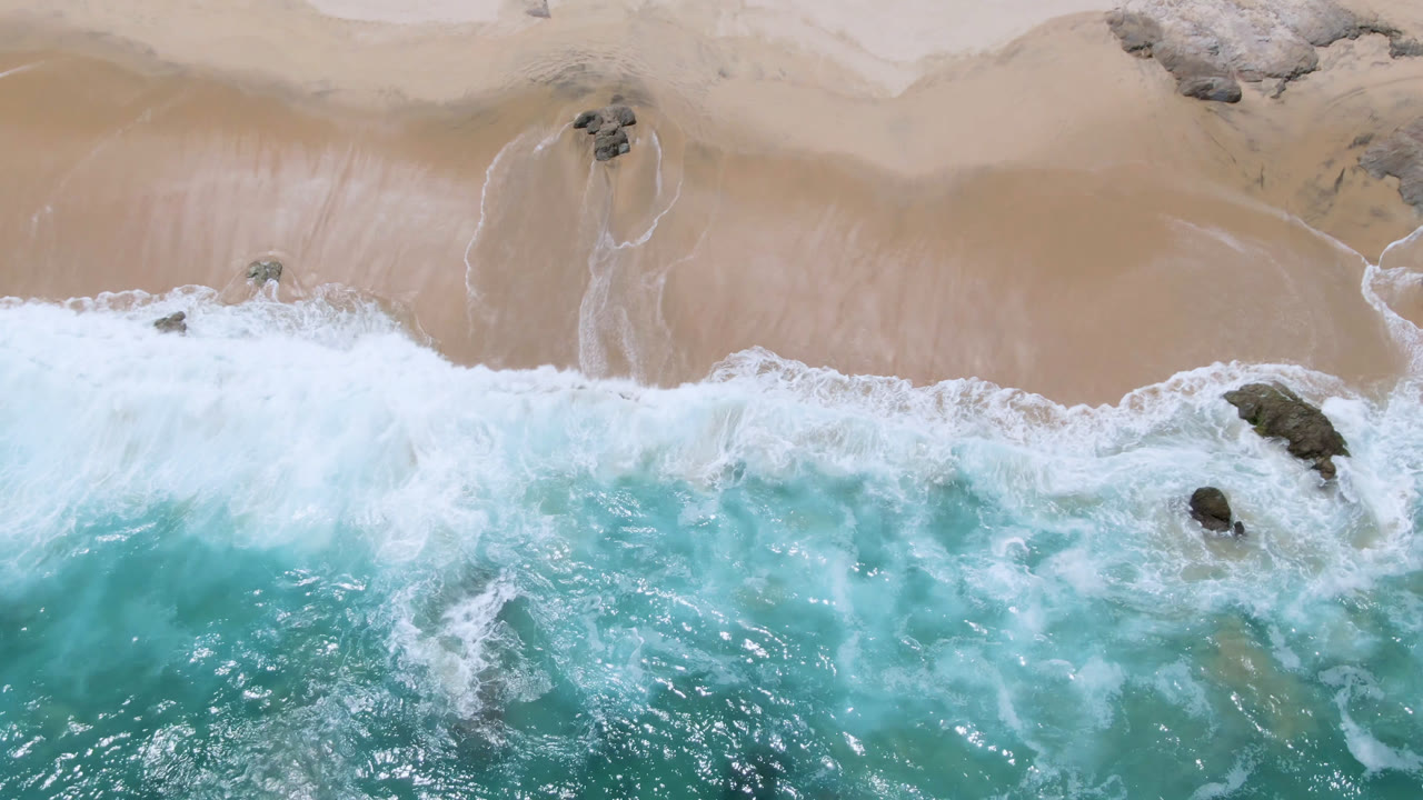 Aerial frame from Laguna Beach, Baja California Sur showing a broader band of white surf spreading over the sand around dark rocks, with no visible people.