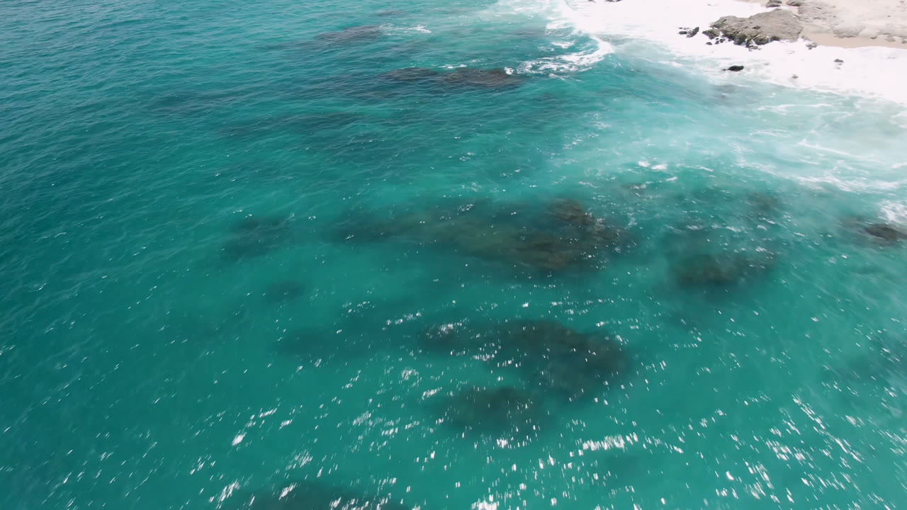 Overhead view of turquoise water at Laguna Beach, Baja California Sur, with dark submerged rocks below the surface, white surf at the upper shoreline edge, and no visible people.