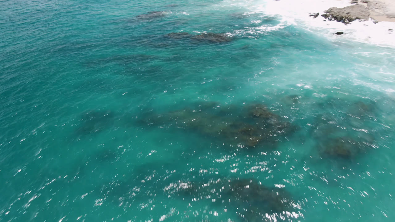 Aerial frame over Laguna Beach, Baja California Sur, showing teal water, scattered dark rock patches under the surface, brighter foam near the top right, and no visible people.