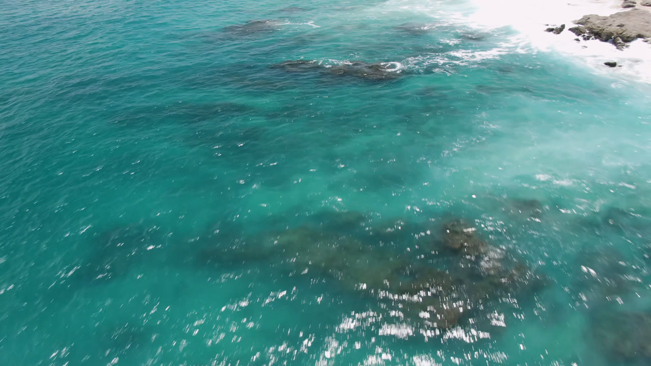 Turquoise ocean with a dark exposed rock cluster near white breaking waves.