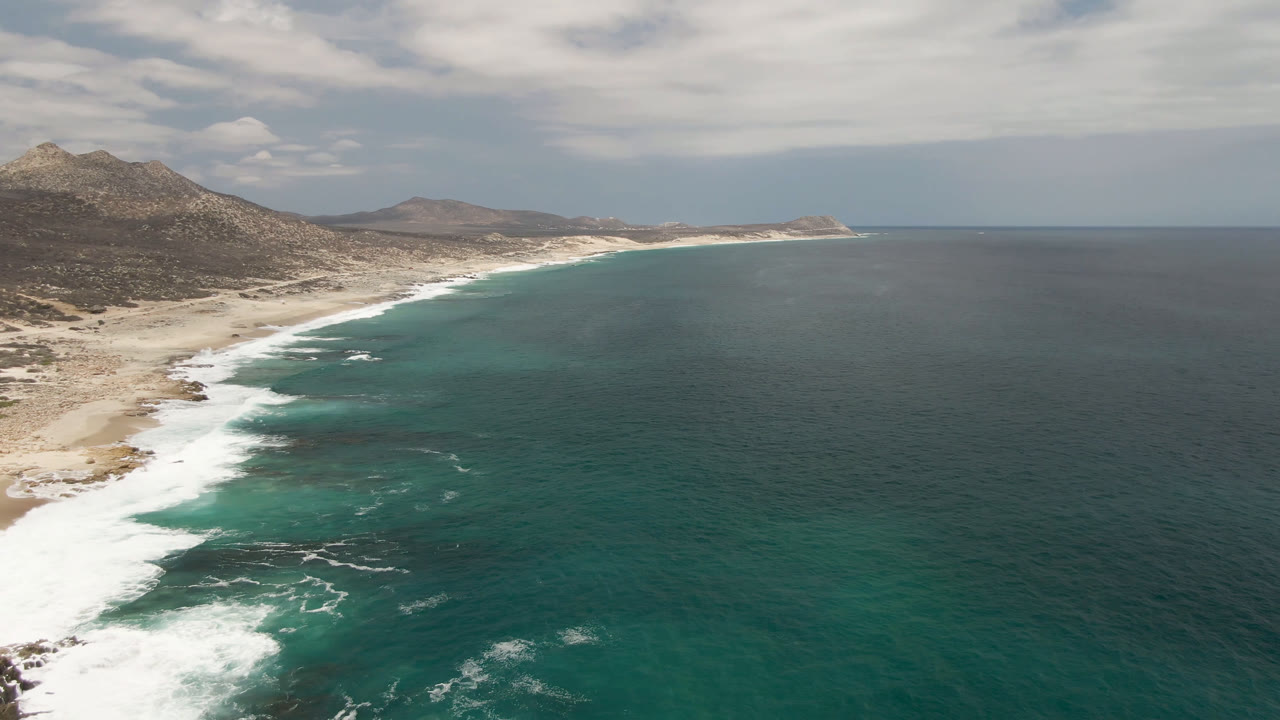 Opening aerial view of Laguna Beach in Baja California Sur with teal water on the right, white surf along the beach, mountains inland, and no visible people.