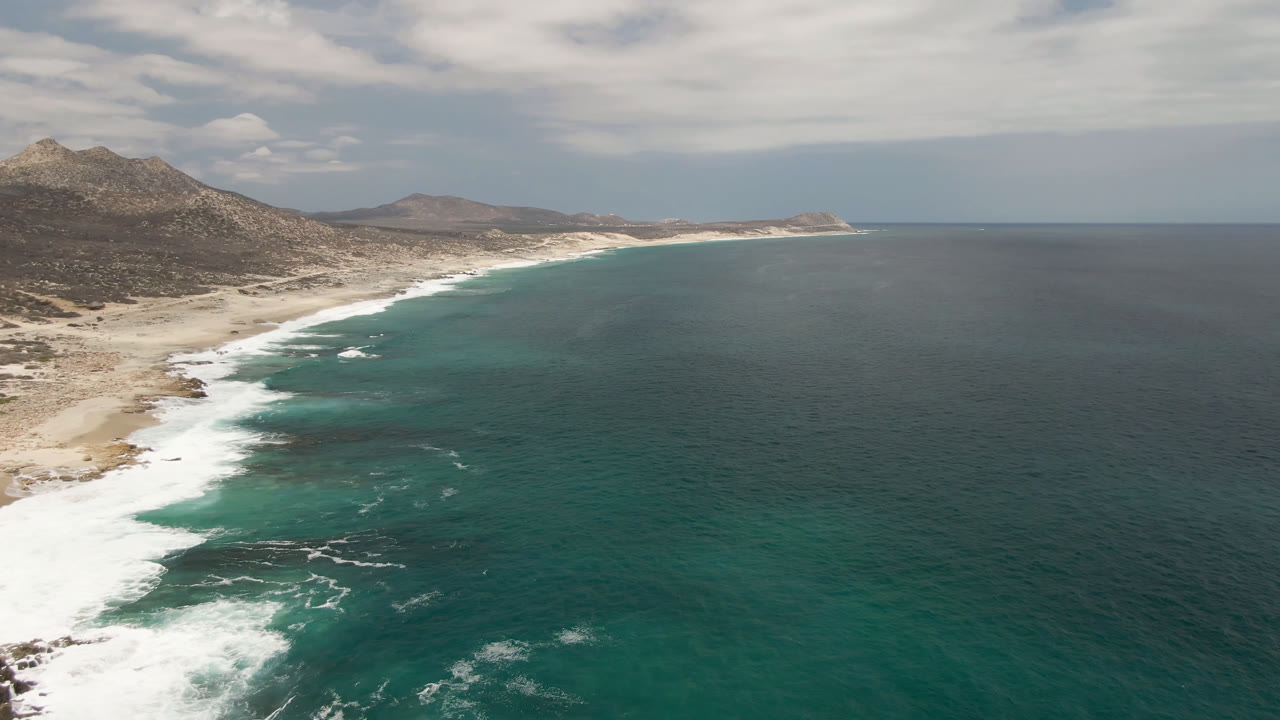 Laguna Beach coastline continues with a long white surf line, pale sand, rocky patches near shore, distant headland, and no visible people.