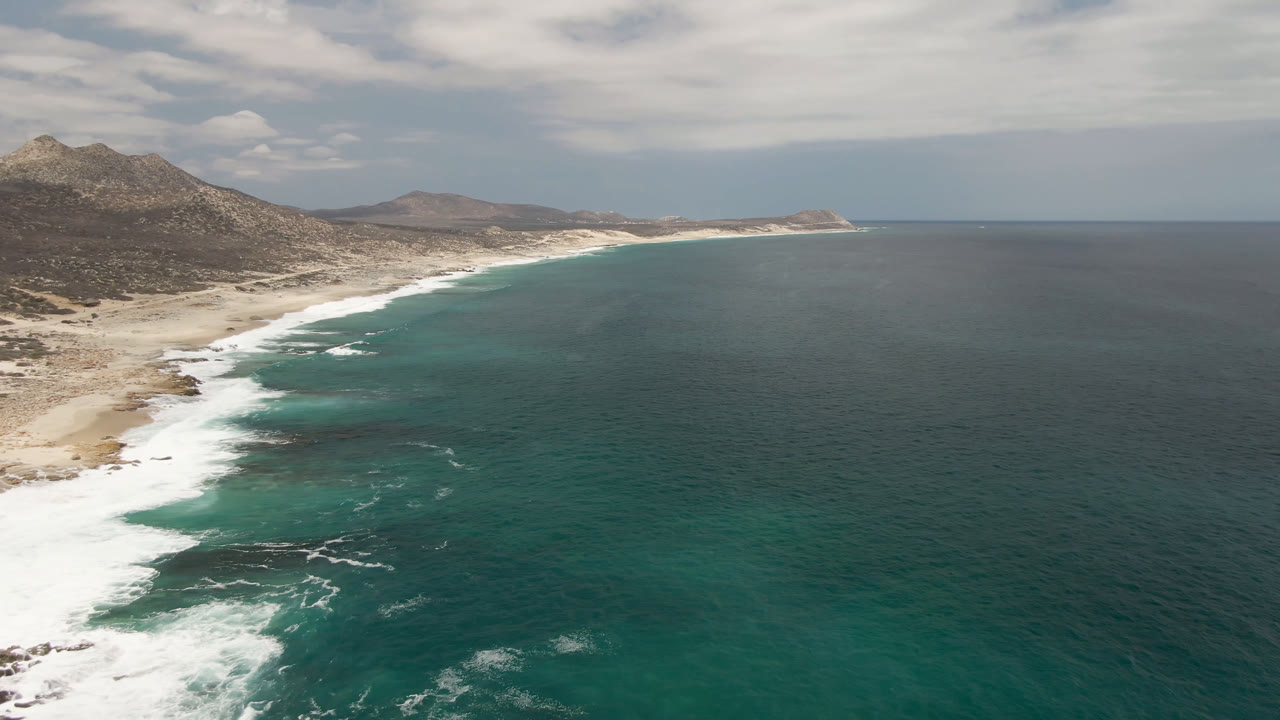 Wide drone view of open ocean beside a curving beach and hills.