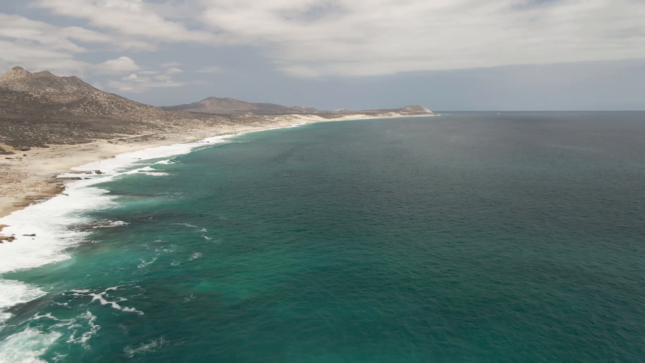 Aerial frame over Laguna Beach in Baja California Sur, with turquoise water in the foreground, white surf along the left shoreline, rocky hills in the background, and no visible people.