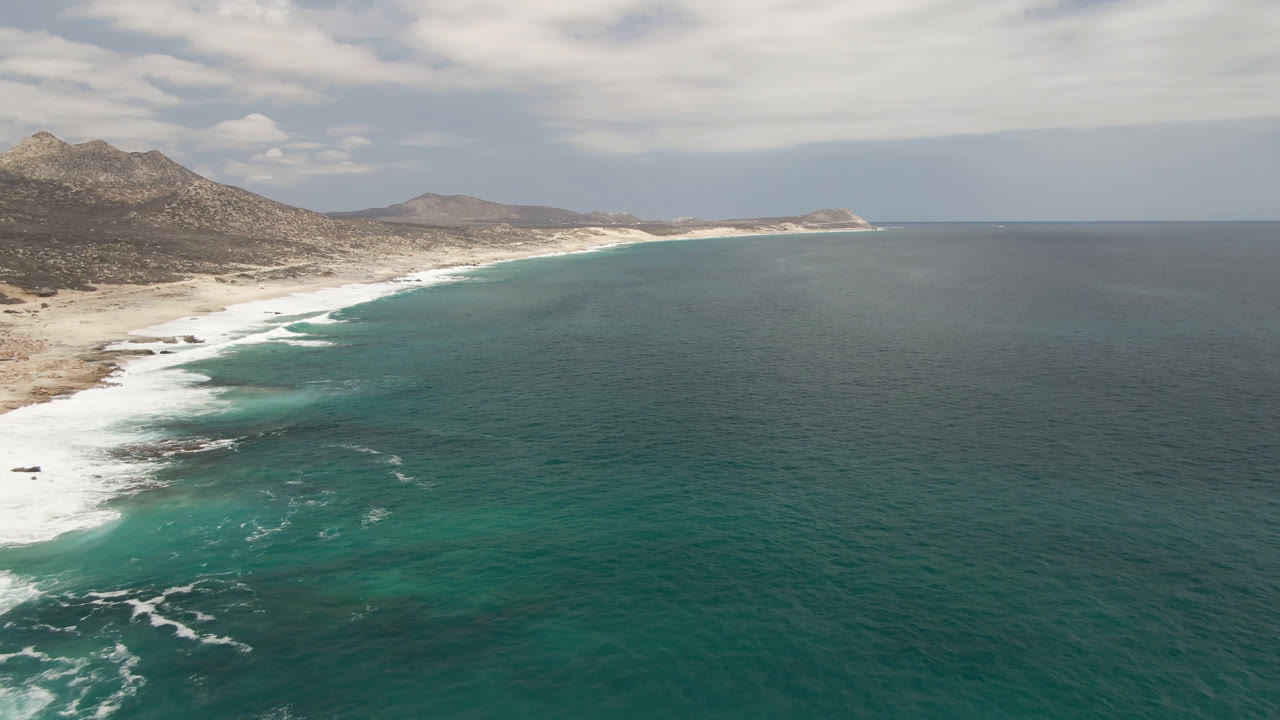 Aerial frame over Laguna Beach, Baja California Sur, showing a long sandy shoreline curving under rocky hills, teal ocean spreading to the right, and no visible people.