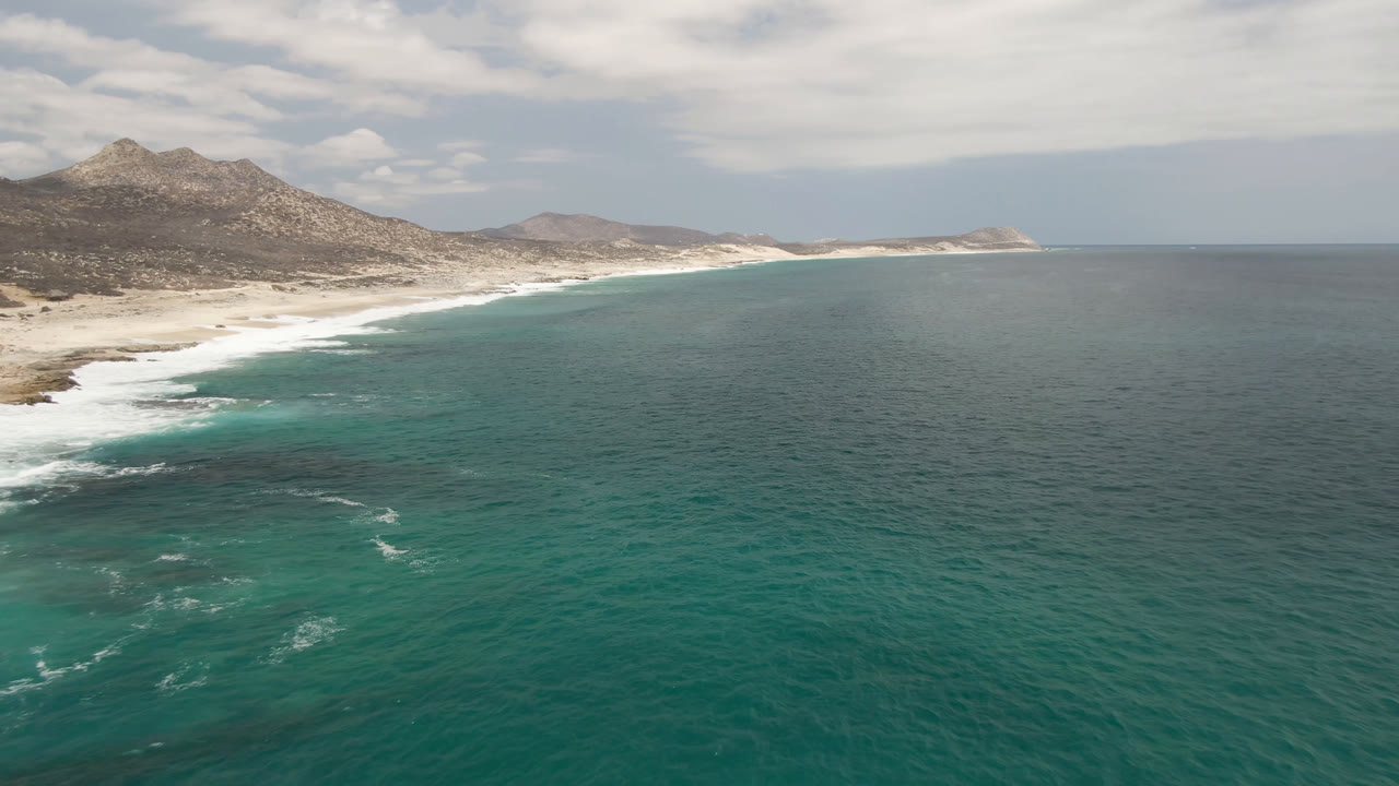 Opening aerial frame of Laguna Beach in Baja California Sur, with teal water in the foreground, a pale curving beach to the left, rocky mountains behind it, cloudy daylight above, and no visible people.