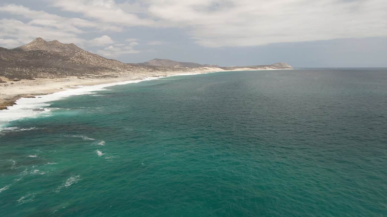 Aerial view at 0:01 over Laguna Beach, Baja California Sur, where thin white wave streaks cross the teal ocean and the sandy shore runs below brown mountains with no visible people.