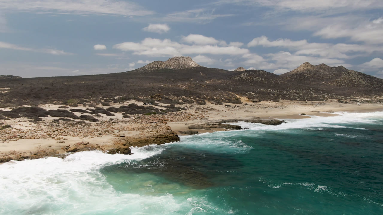 Aerial frame over Laguna Beach in Baja California Sur, Mexico, with turquoise surf breaking over dark rocks, pale sand beyond, low mountains in the background, and no visible people.