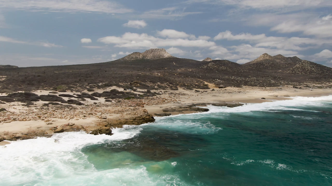 Laguna Beach coastline from above, showing white water spread wider across the left foreground, a rocky tan point at center, distant scrub hills, mountains, and no visible people.