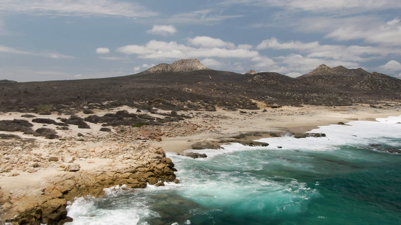 Aerial view of Laguna Beach, Baja California Sur, with white surf curling around tan rocks, pale sand beyond, dark scrubland, low hills, scattered clouds, and no visible people.