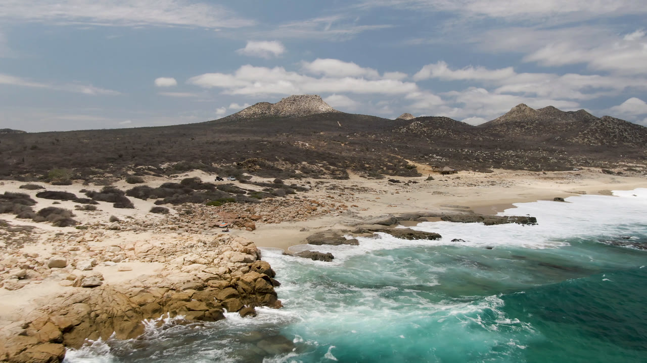 Laguna Beach, Baja California Sur shoreline with foamy water pushing into a rocky cove, a wide strip of pale sand, scrub-covered ground, distant hills, and no visible people.